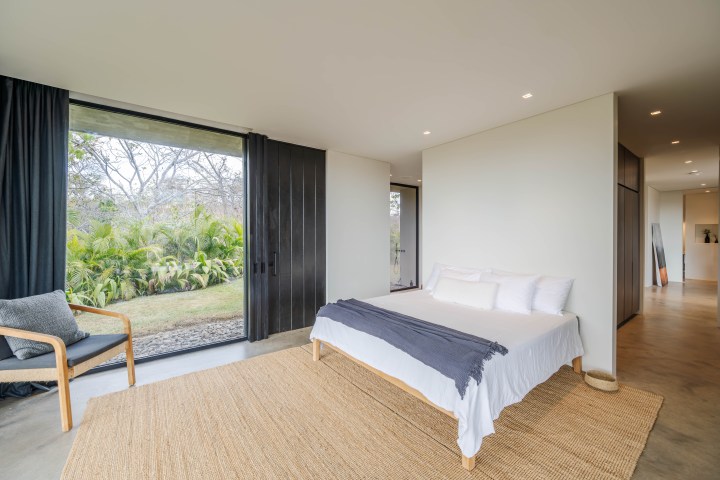 Minimalist bedroom with large window, bed, chair, rug, and hallway view.
