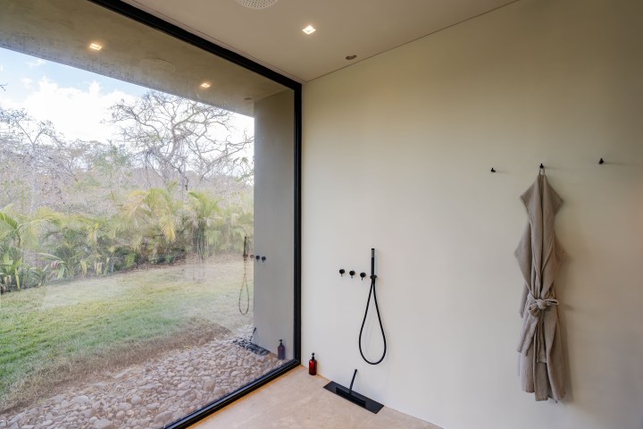 Minimalist bathroom with large glass window showing outdoor greenery, wall-mounted shower, and hanging towels.