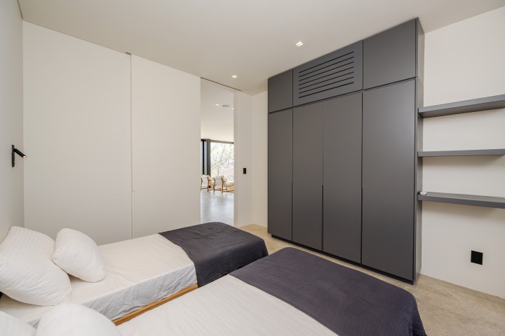 Minimalist bedroom with twin beds, dark wardrobe, and open sliding door to a bright living room.