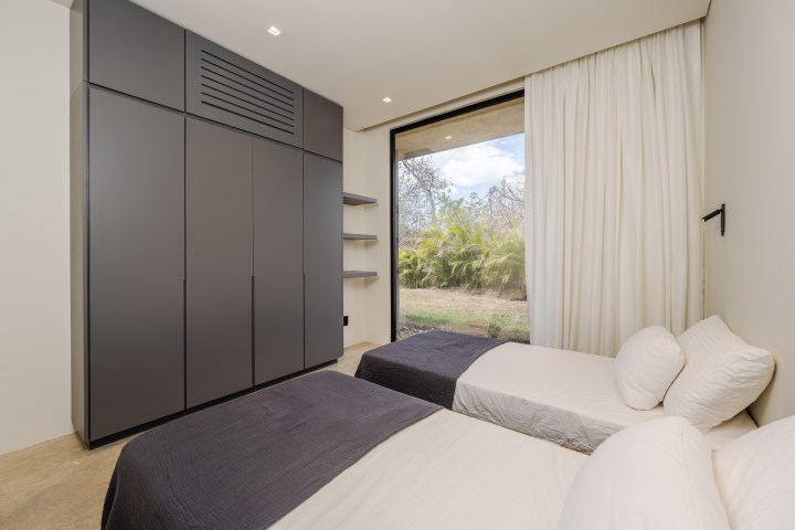 Minimalist bedroom with two single beds, gray wardrobe, and large window with garden view.