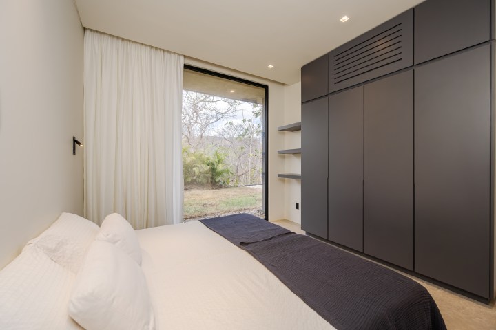Modern bedroom with white bed, gray wardrobe, large window, and view of trees.