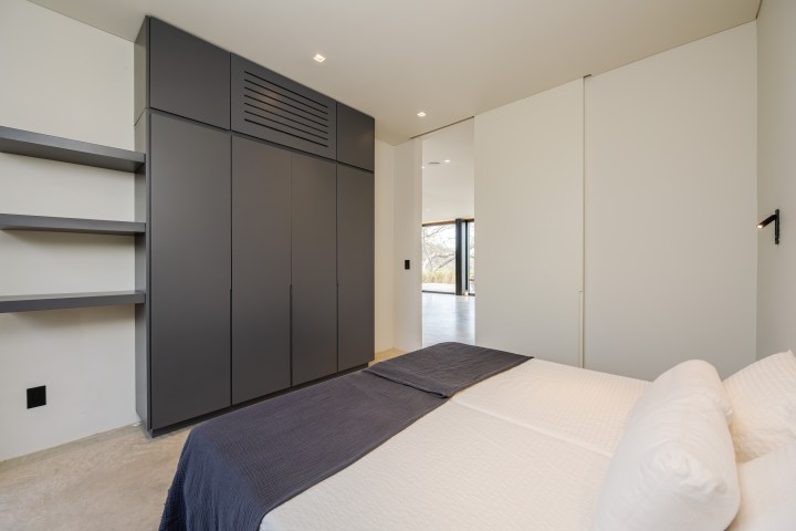 Minimalist bedroom with a double bed, built-in wardrobe, shelves, and an open doorway.