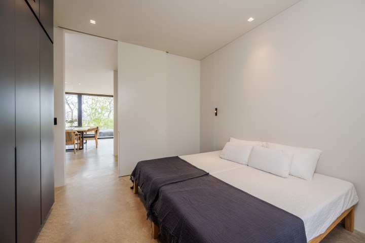 Minimalist bedroom with twin bed, dark wardrobe, and open doorway to a bright room with a table.