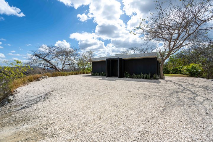 Modern black building surrounded by gravel and sparse trees under a partly cloudy sky.