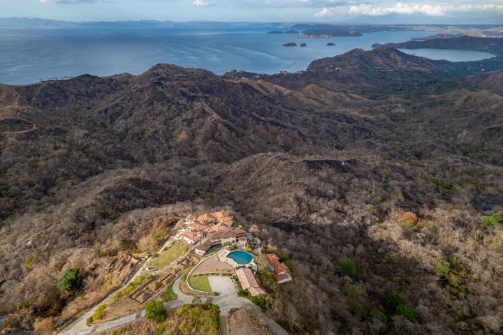 Aerial view of a hillside mansion with a pool, surrounded by dry landscape, overlooking distant ocean and islands.