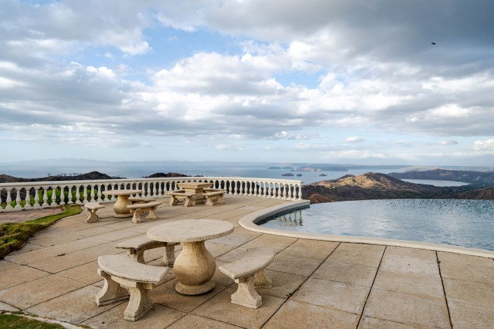 Stone patio with tables, infinity pool, and ocean view under cloudy sky.