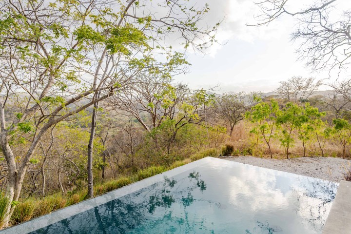 Infinity pool reflecting trees and cloudy sky, surrounded by sparse greenery and distant hills.