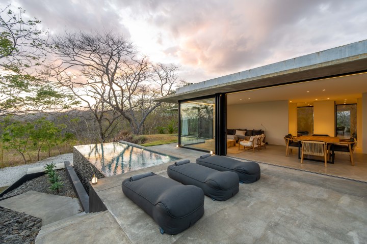 Modern patio with loungers, infinity pool, and open living space at sunset.