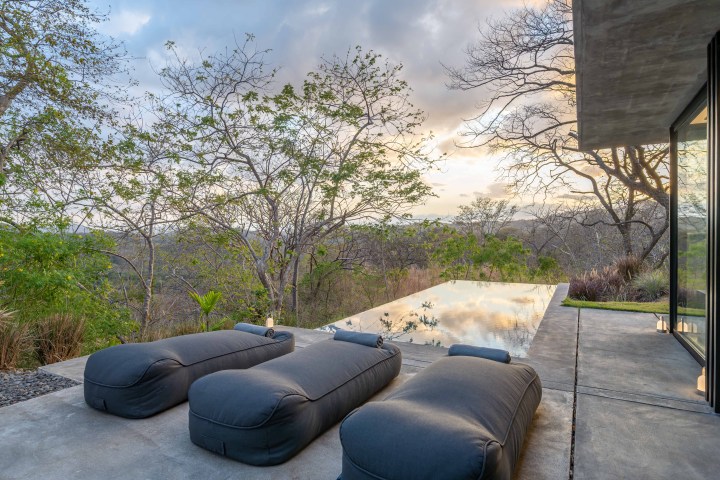 Outdoor loungers by an infinity pool overlooking a scenic forest view at sunset.