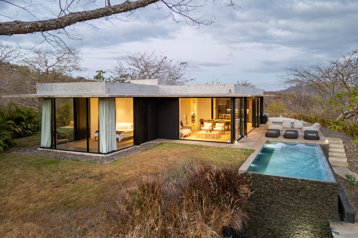 Modern house with large glass windows, patio seating, and an infinity pool, surrounded by trees and grass.