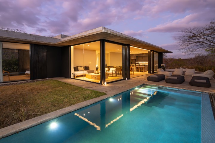 Modern house with large windows, lit interior, pool, and outdoor seating at dusk.