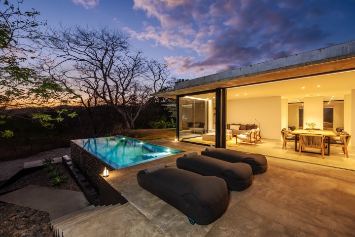 Modern house with glass walls, pool, and seating area at sunset, surrounded by trees.