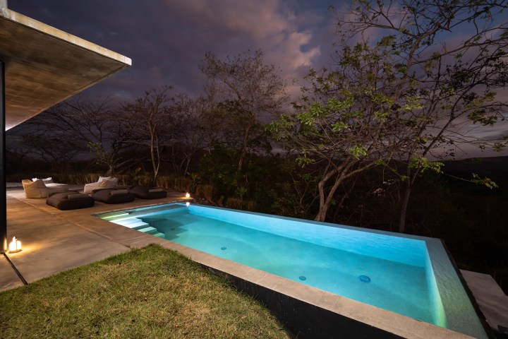 Modern pool at night with lounge chairs, surrounded by trees and a concrete patio.