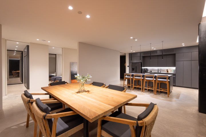 Modern kitchen with wooden dining table, chairs, and dark cabinets, under ambient lighting.