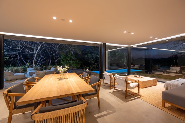 Modern living room with large windows, wooden furniture, and view of outdoor seating at dusk.