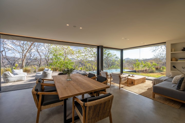 Spacious living room with large windows, wooden table, and outdoor view of trees and lawn.