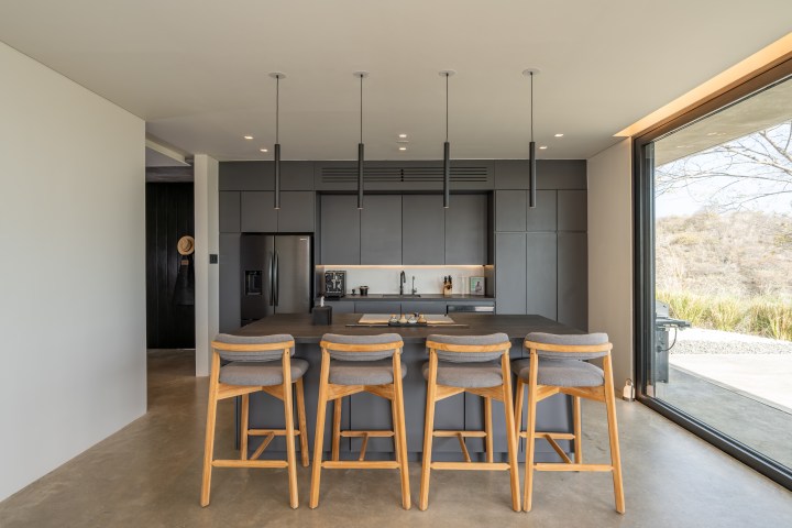 Modern kitchen with gray cabinets, island, four chairs, hanging lights, and large window view.