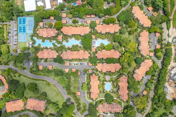 Aerial view of buildings with red roofs, green trees, winding pool, and tennis courts.