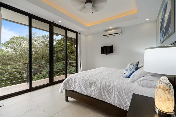 Modern bedroom with large windows, white bedding, and forest view.