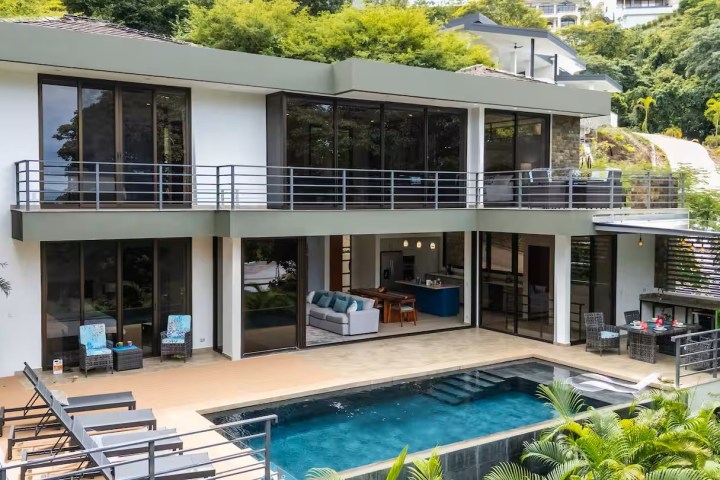 Modern two-story house with large glass windows and a pool in a lush setting.