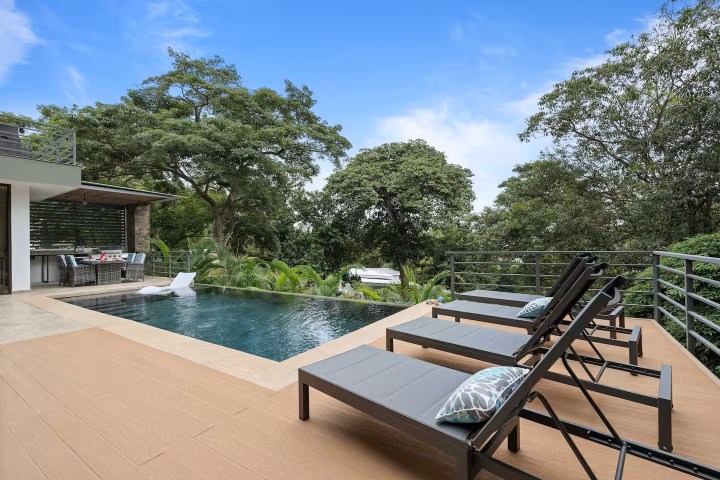 Modern patio with pool, sun loungers, and an outdoor dining area, surrounded by trees.