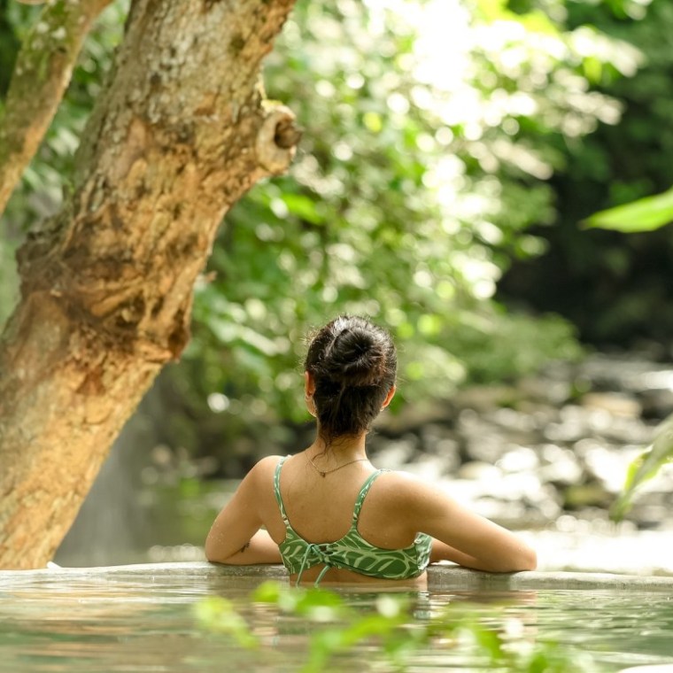 Woman in a swimsuit relaxes in a woodland hot spring, surrounded by lush greenery.