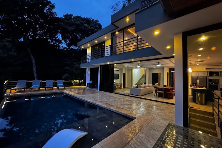 Modern two-story house with pool at dusk, interior lights on.