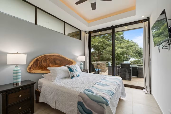 Modern bedroom with large windows, wooden headboard, and balcony view of trees.