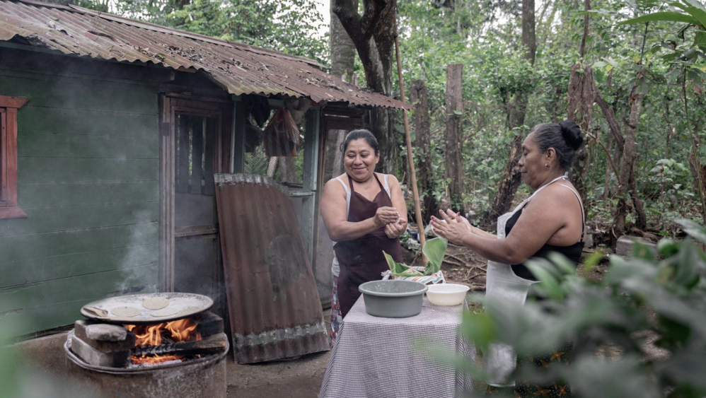 Two women making tortillas outdoors beside a rustic wooden house with a fire stove.