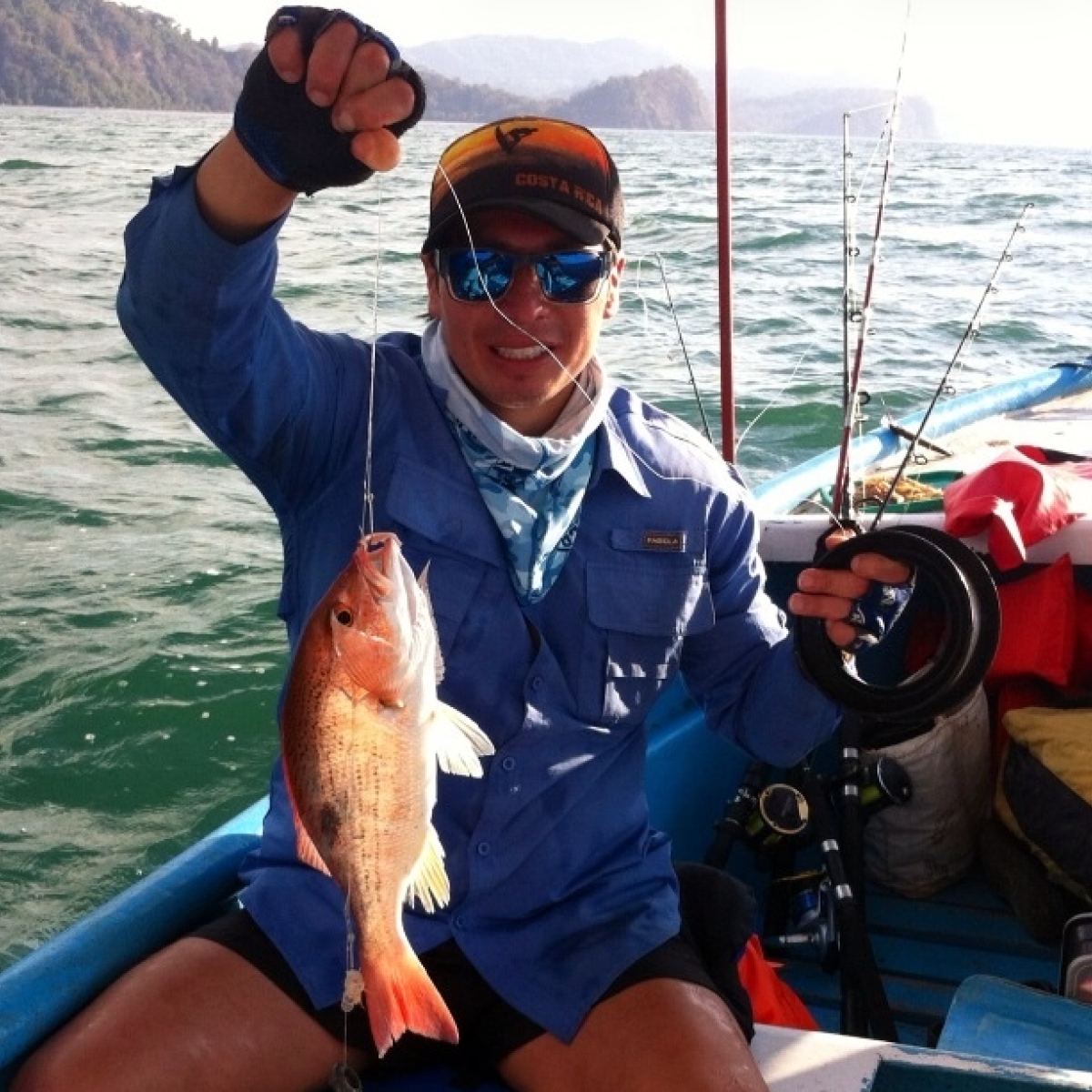 Person in blue shirt holding a fish on a boat in a coastal area.