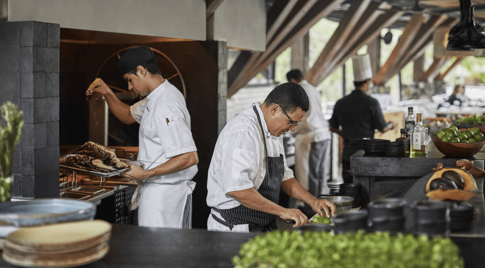 Chefs in a busy restaurant kitchen preparing food, one slicing vegetables and another handling grilled meat.
