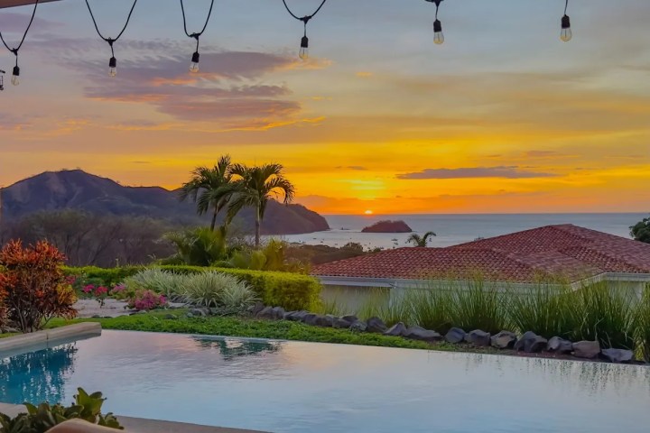 Sunset view over pool, ocean, and mountains with string lights and palm trees.