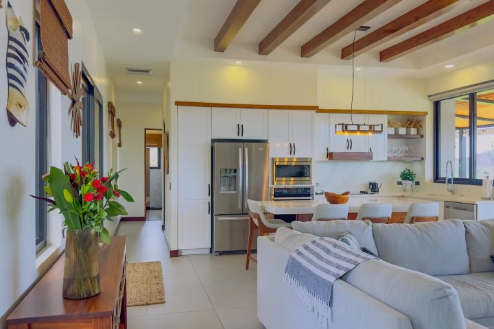 Modern kitchen and living area with white cabinets, wood beams, and gray couch.