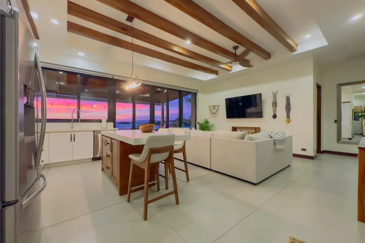 Modern open-plan kitchen and living room with sunset view through large windows.