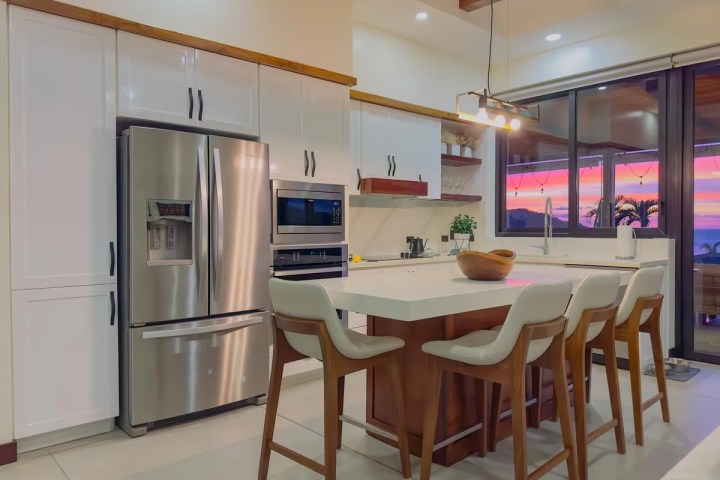 Modern kitchen with white cabinets, island, stainless steel appliances, and sunset view through large windows.