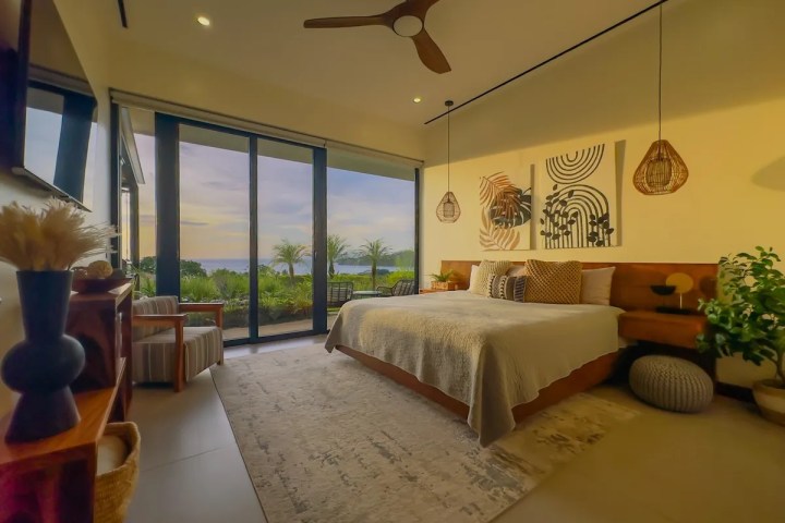 Modern bedroom with ocean view, large windows, cozy bed, plants, and abstract wall art.