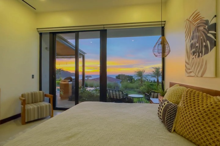 Room with a bed, large window view of sunset, chairs, and patio.