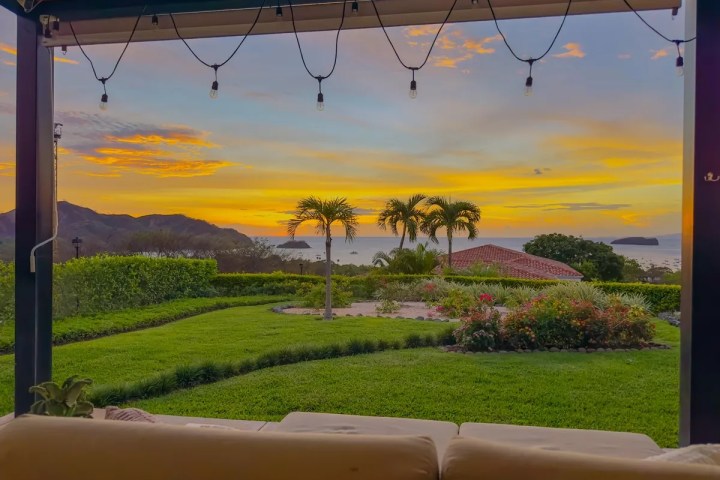 Sunset view from patio with lights, palm trees, and ocean in background.