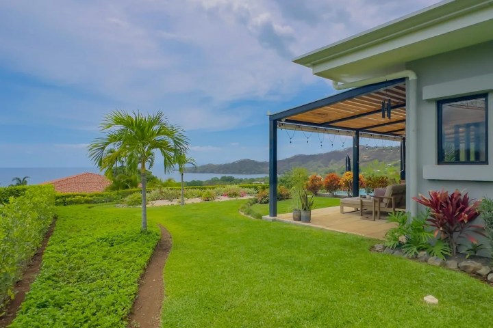 Covered patio with seating, overlooking ocean and garden with palm trees.