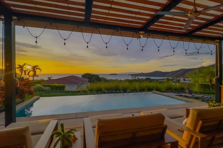 Infinity pool and patio with ocean view at sunset under pergola with hanging lights.