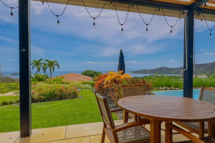 Patio with wooden table, chairs, ocean view, and garden in the background.
