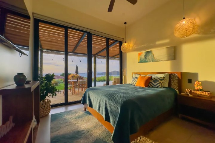 Bedroom with teal bedspread, patio doors, two hanging lights, and outdoor view at sunset.