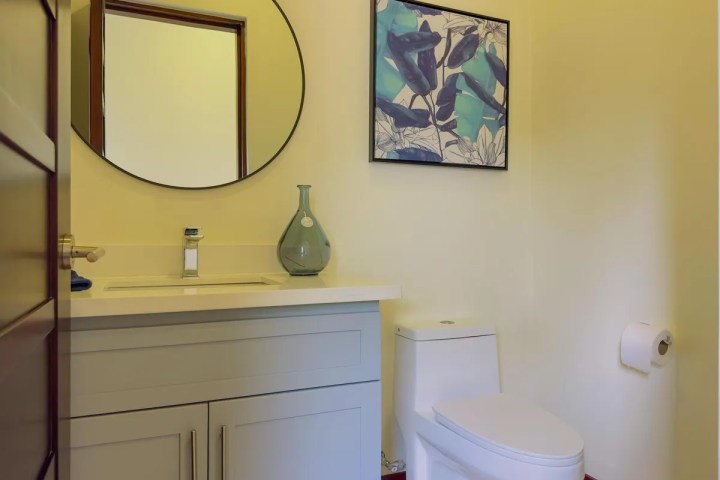 Bathroom with round mirror, vanity, toilet, wall art, and a glass vase on the counter.