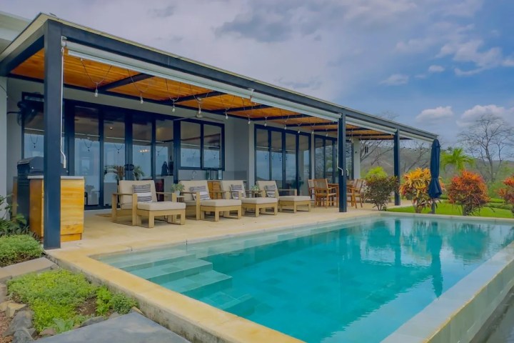 Modern house with patio and pool, surrounded by greenery under a cloudy sky.