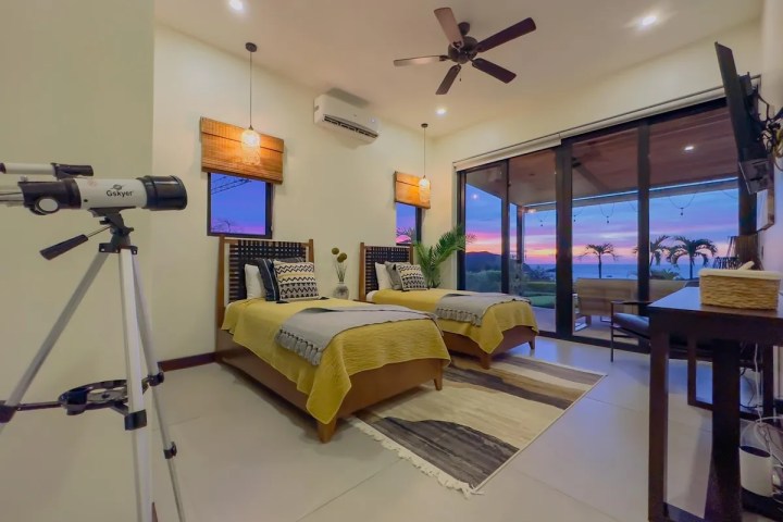 Bedroom with twin beds, telescope, ceiling fan, and ocean view through large windows at sunset.