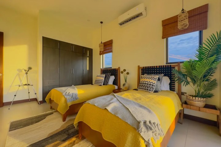 Room with two single beds, yellow bedding, large wardrobe, telescope, and potted plant.