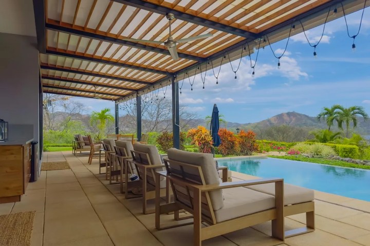 Covered patio with lounge chairs overlooking pool, tropical plants, and mountains.
