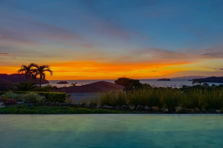 Sunset over a coastal landscape with palm trees and a tiled roof.