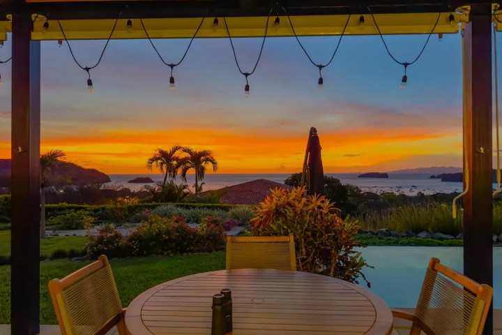 Outdoor patio with wooden table overlooking a sunset view of the ocean and palm trees.