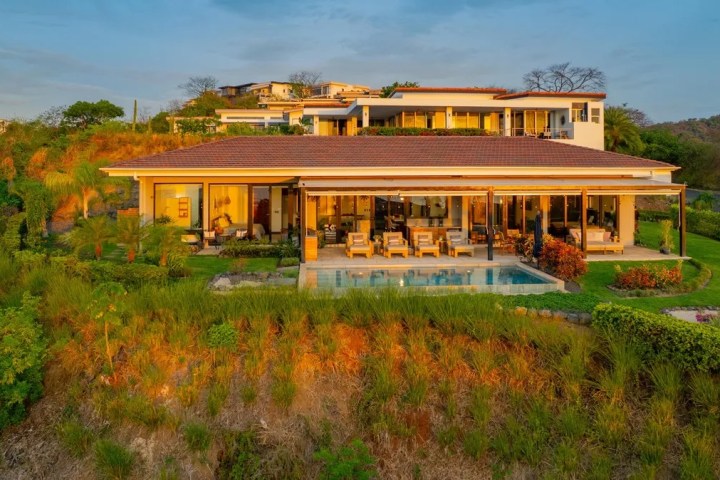 A luxurious modern house with a pool, surrounded by lush greenery and set on a hillside.